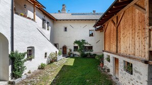 A courtyard with a stone pathway, a grassy area, and a building with wooden beams and white walls. - Apartment 'Schloss Gravetsch Castel' with Mountain View and Shared Garden (Villandro)
