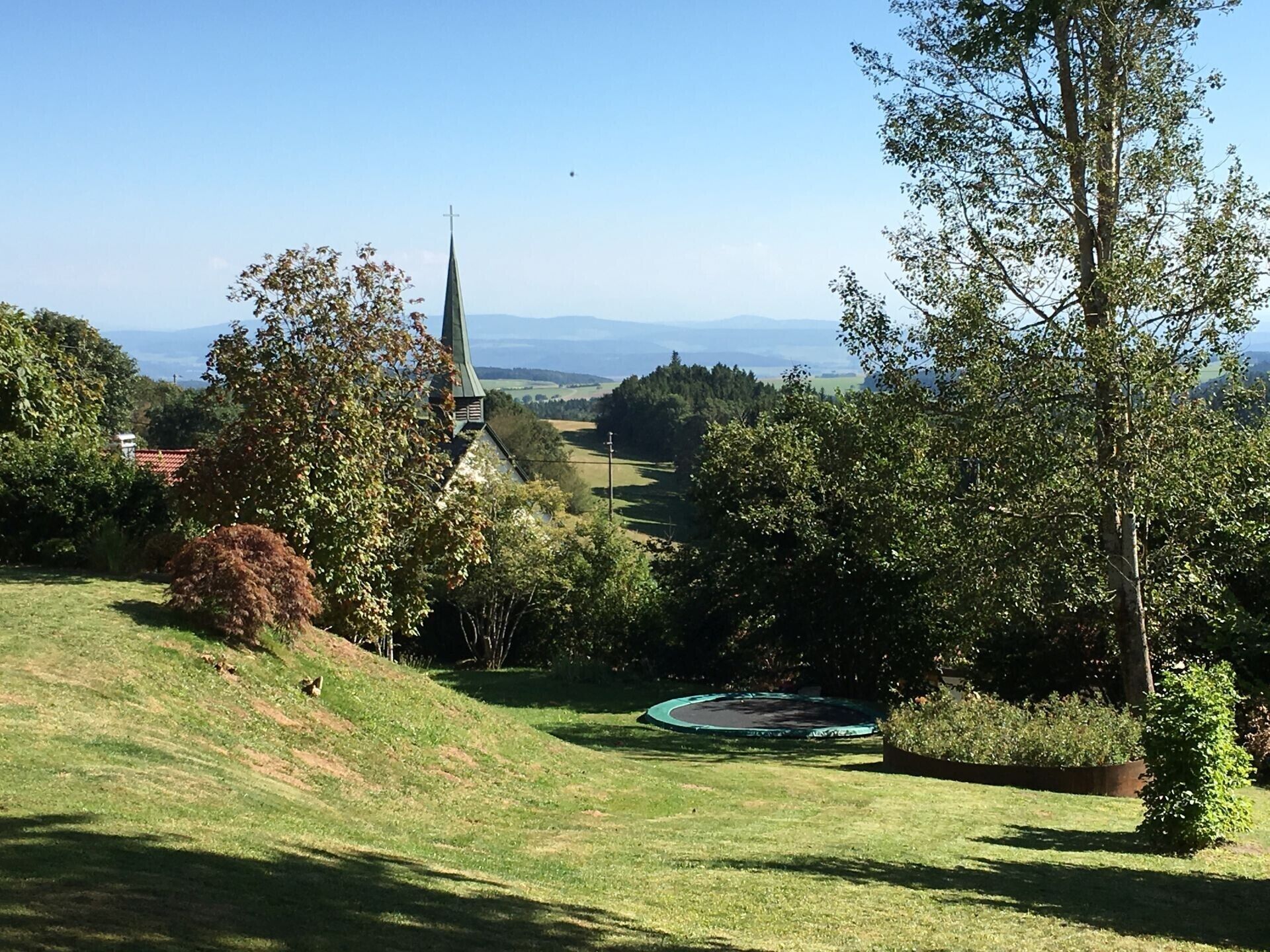 Une église dotée d’un clocher, entourée d’arbres et bordée d’une colline verdoyante.