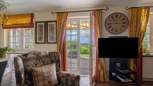 A living room with a sofa, a television, a large wall clock, and a view through a glass door.