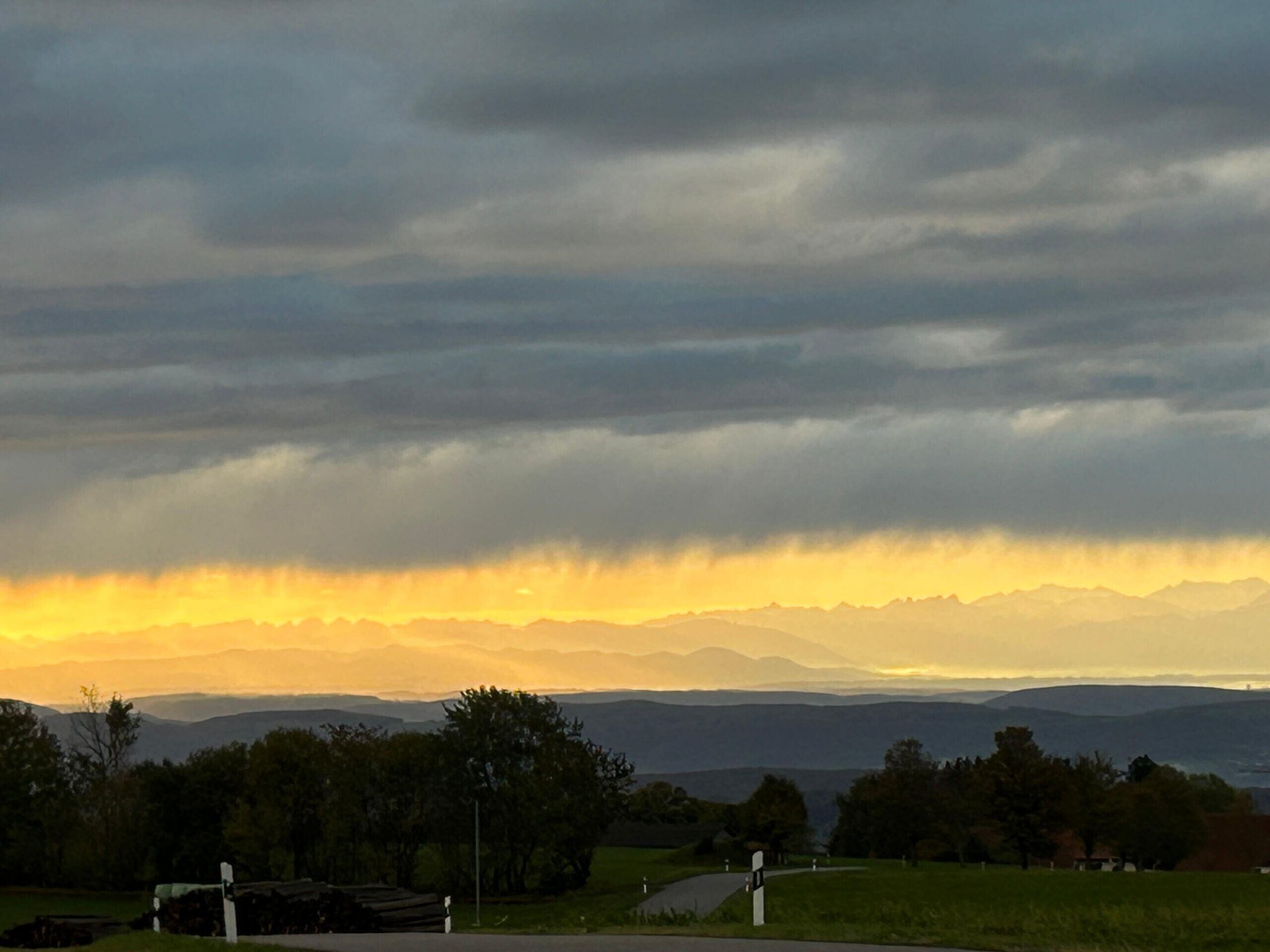 A rural landscape with a road, trees, and distant mountains under a cloudy sky.