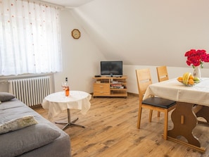 Ein gemütliches Zimmer mit einer Couch, einem Esstisch mit Stühlen, einem Fernseher auf einem Holzgestell, einem kleinen runden Tisch mit Flasche und Glas sowie einem Heizkörper.