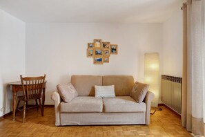 A living room with a beige sofa, a wooden chair, a wall-mounted picture frame, a standing lamp, and a radiator.