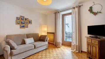 Ett vardagsrum med en soffa, en tv, ett träskåp, en väggmonterad lampa och ett fönster med gardiner.