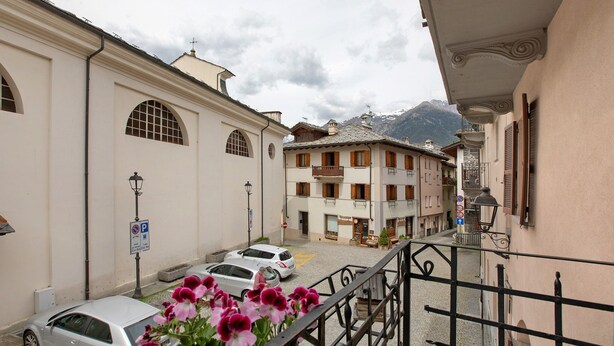 Une rue étroite bordée de voitures garées, un immeuble avec un balcon et une montagne en arrière-plan.