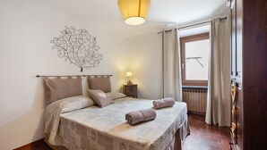 Une chambre à coucher comprenant un lit, une table de chevet, un tableau décoratif au mur, une fenêtre avec des rideaux et une commode en bois.
