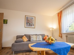 Una sala de estar con un sofá gris, un cuadro floral enmarco, un frutero y una mesa con mantel azul.