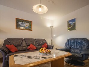 A living room with a leather sofa, a dining table with fruit, a floor lamp, and two framed pictures on the wall.