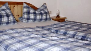 Un lit avec une couvre-lit à carreaux bleus et blancs, une tête de lit en bois et deux oreillers. Un tableau représentant une rose est accroché au mur, et une petite table avec un verre et une plante se trouve à côté du lit.
