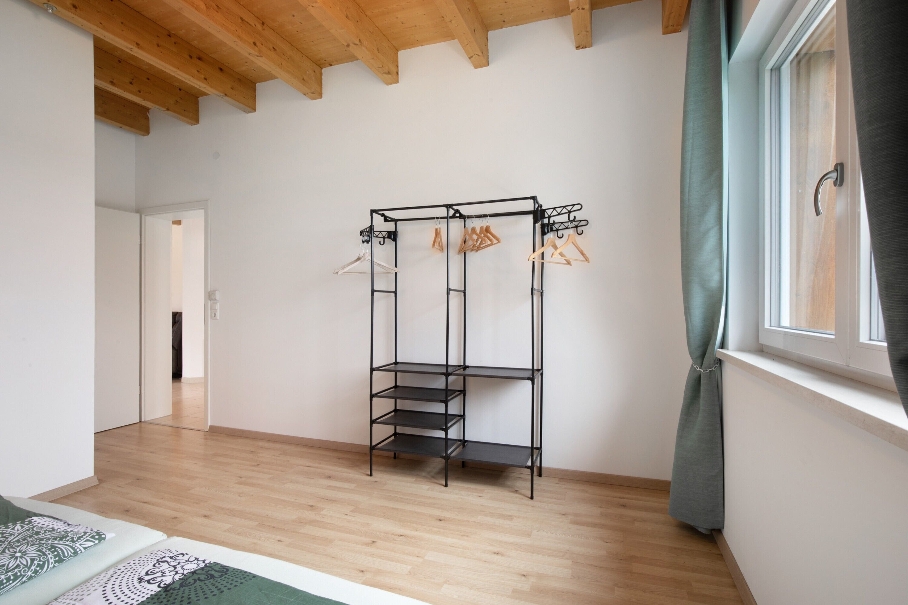 Une pièce avec un plafond en bois, une étagère en métal noir où sont pliés des vêtements, une fenêtre avec un rideau et un plancher en bois.