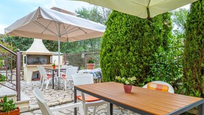 An outdoor patio with stone flooring, white chairs, a wooden table, a grill, and a large umbrella.