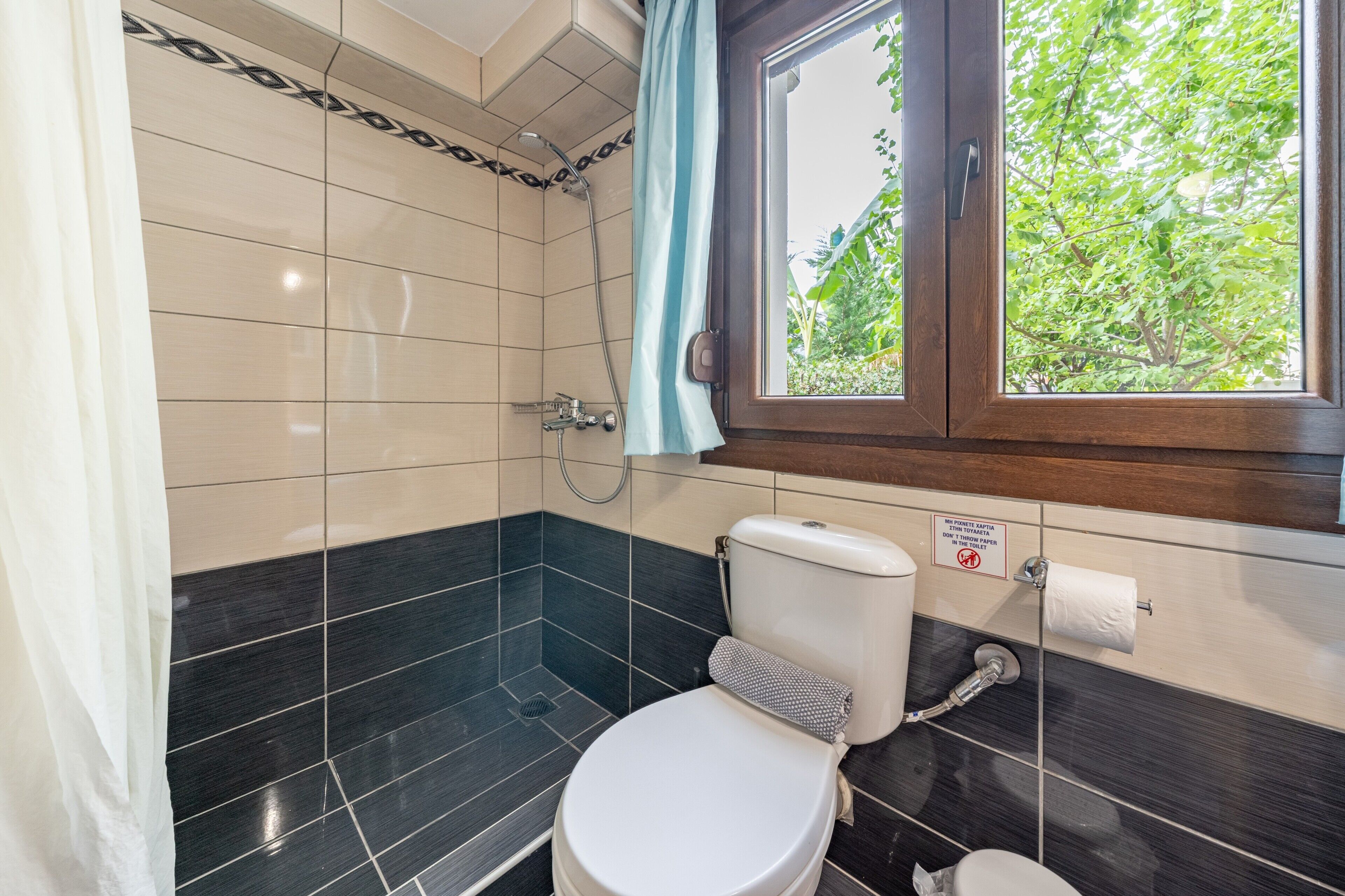 A bathroom with a toilet, a shower, and a window with a view of greenery.