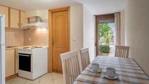 A compact kitchen with a stove, oven, and wooden cabinets. There is a dining table with two chairs and a checkered tablecloth. A door leads to a balcony with a view of greenery.