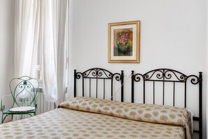 A bed with a patterned bedspread, a green metal chair, a framed picture on the wall, and a window with white curtains. - Room 'Camera 43' with Wi-Fi and Air Conditioning (Verona)