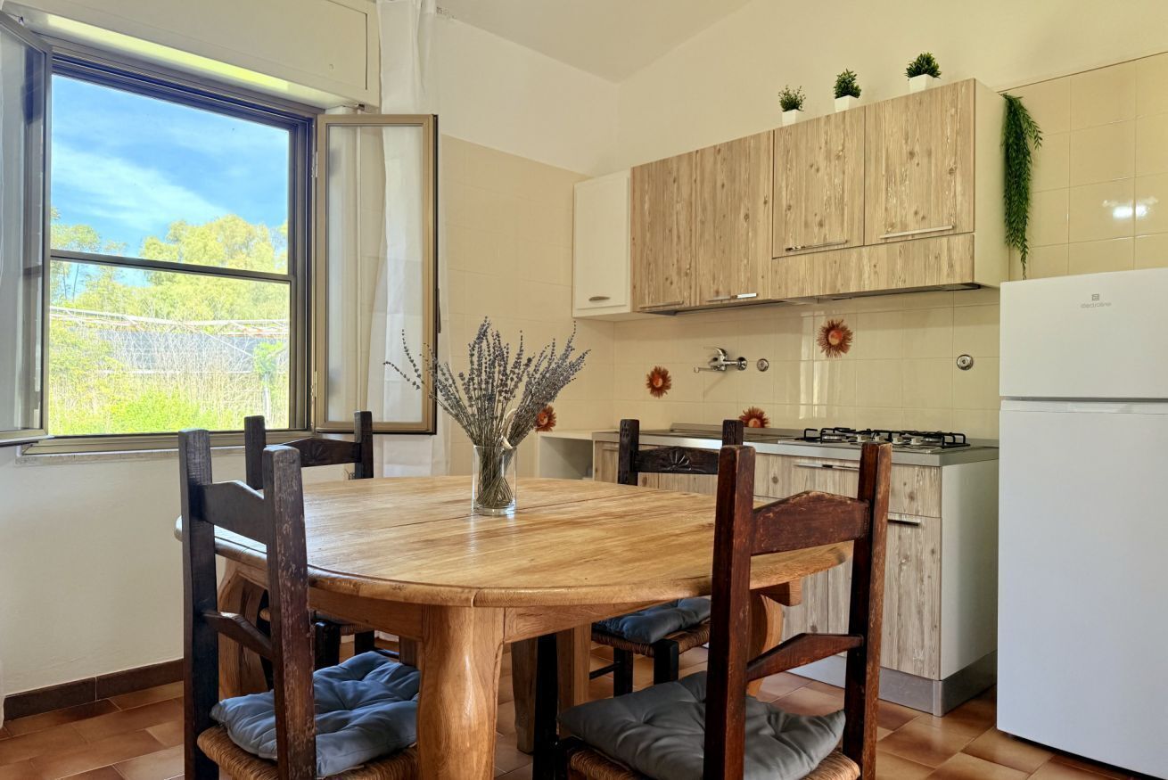 Une cuisine avec des meubles en bois, une table à manger avec des chaises, un réfrigérateur et une fenêtre donnant sur des arbres.