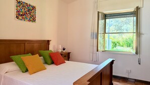 A bedroom with a wooden headboard, a bed with white bedding and colorful pillows, a nightstand with a lamp, and a window with curtains.