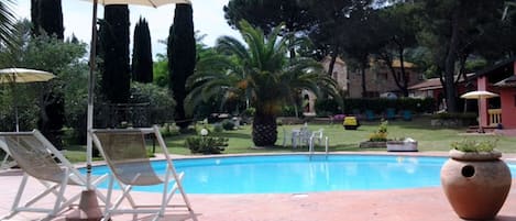 Una piscina con sillones y sombrillas, rodeada de vegetación y edificios.