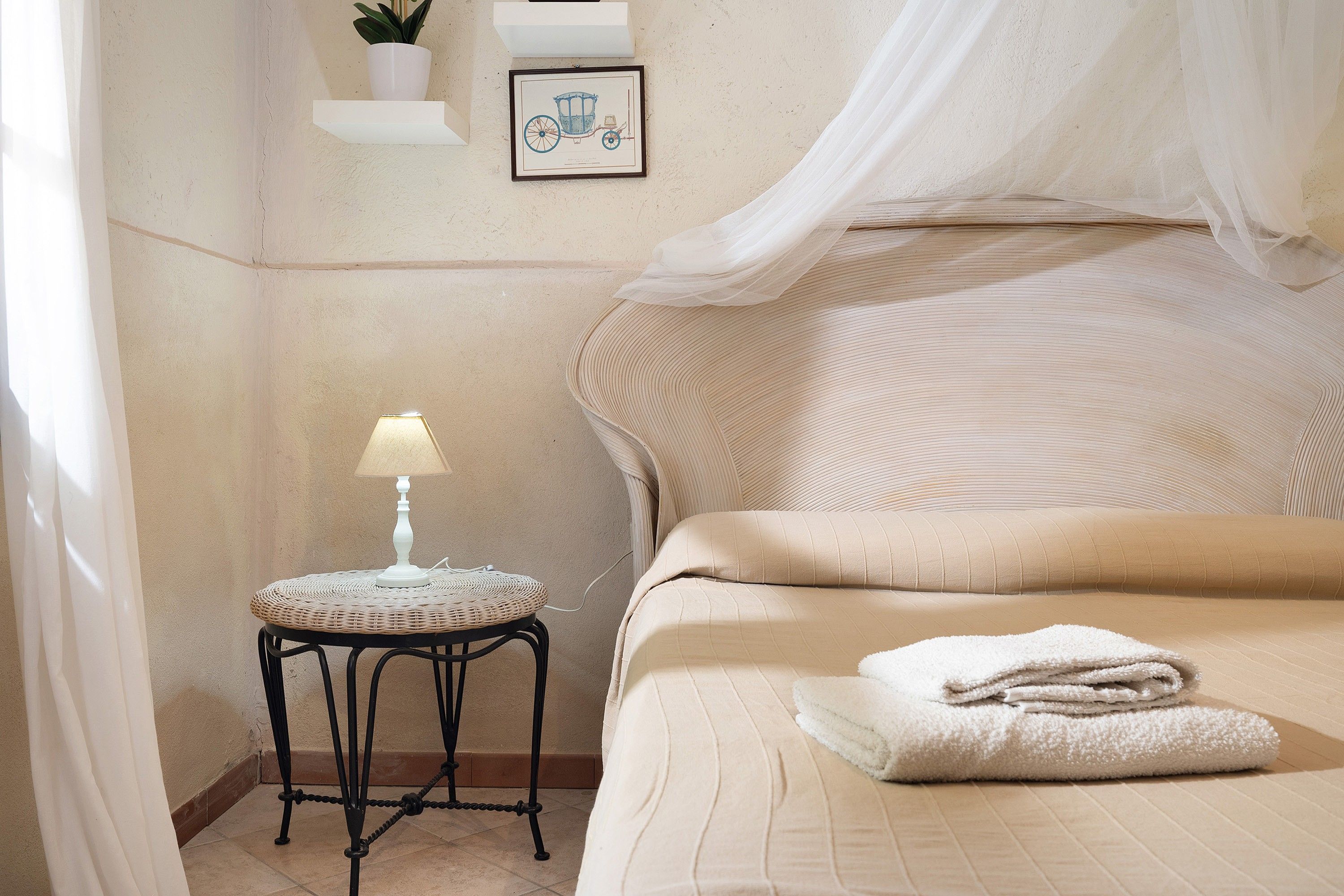 A bedroom with a bed, a bedside table with a lamp, a wall-mounted shelf with a plant, and a framed picture.