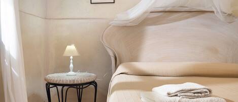 A bedroom with a bed, a bedside table with a lamp, a wall-mounted shelf with a plant, and a framed picture.