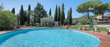 Una piscina con borde de azulejos, rodeada por un jardín con árboles altos y un edificio de piedra al fondo.