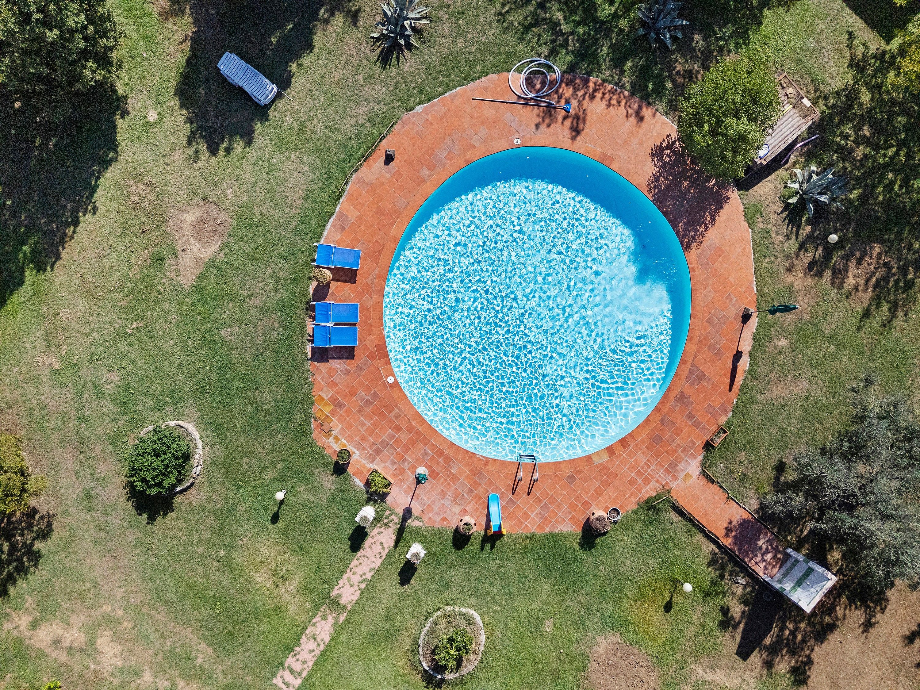 Una vista aerea di una piscina circolare con un bordo piastrellato, panchine e un piccolo giardino.