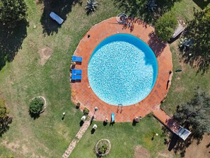 An aerial view of a circular swimming pool with a tiled deck surrounding it, benches, and a small garden area.