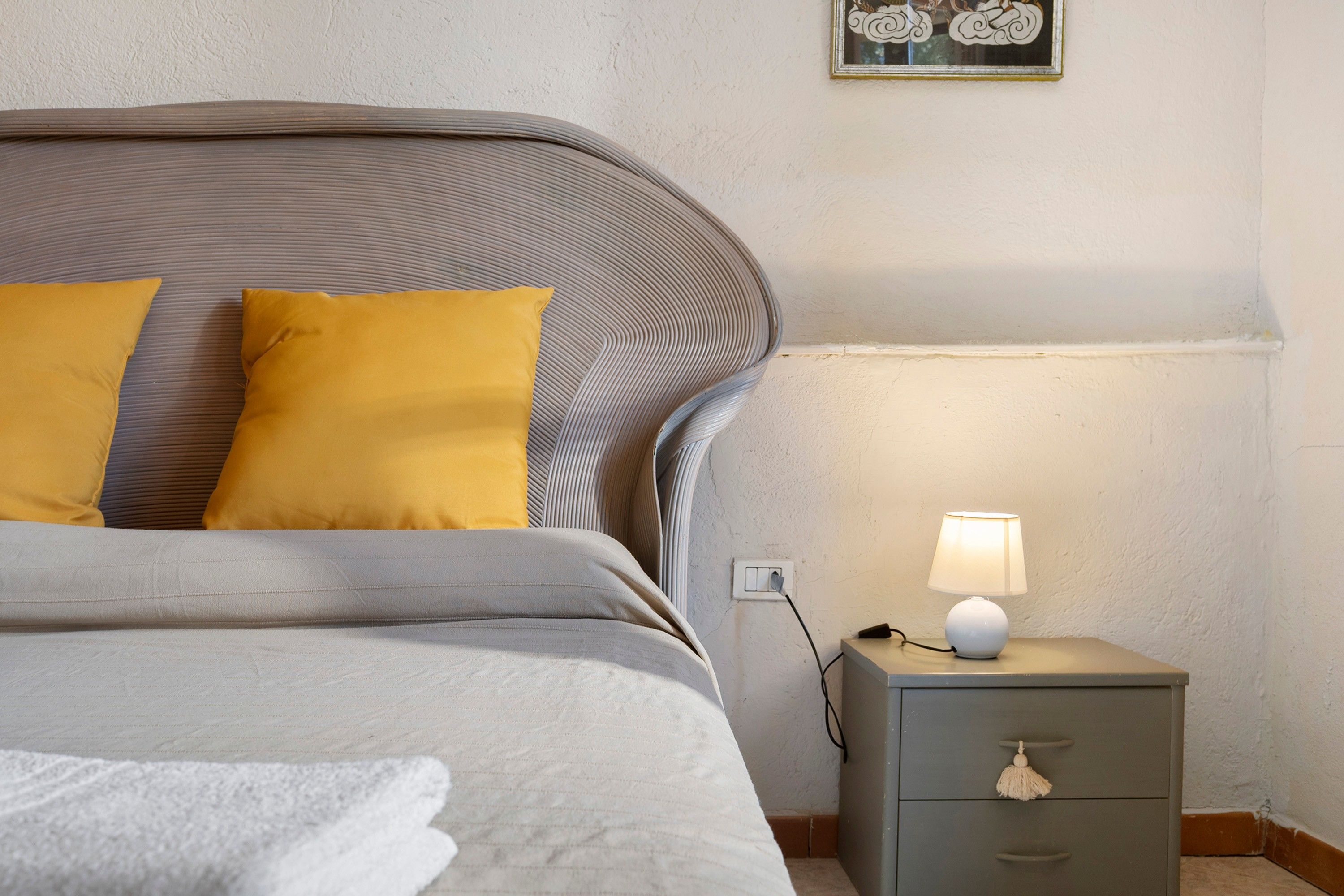Una cama bien hecha con cabecera gris y dos almohadas amarillas, una mesita de noche con lámpara y un pequeño aparador.
