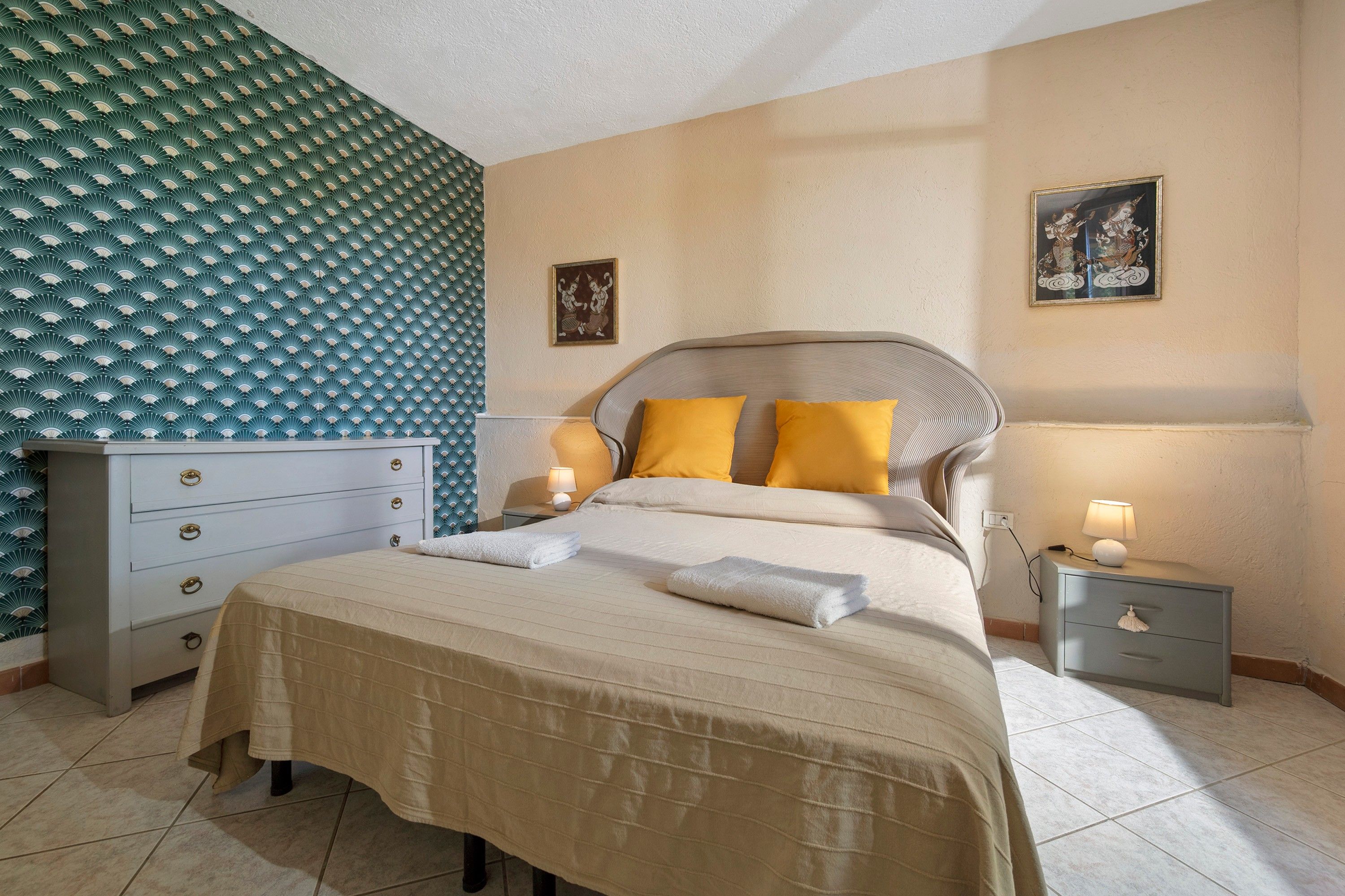 A neatly made bed with a beige bedspread and two pillows, a bedside table with a lamp, a dresser with drawers, and a wall with a patterned wallpaper and two framed pictures.