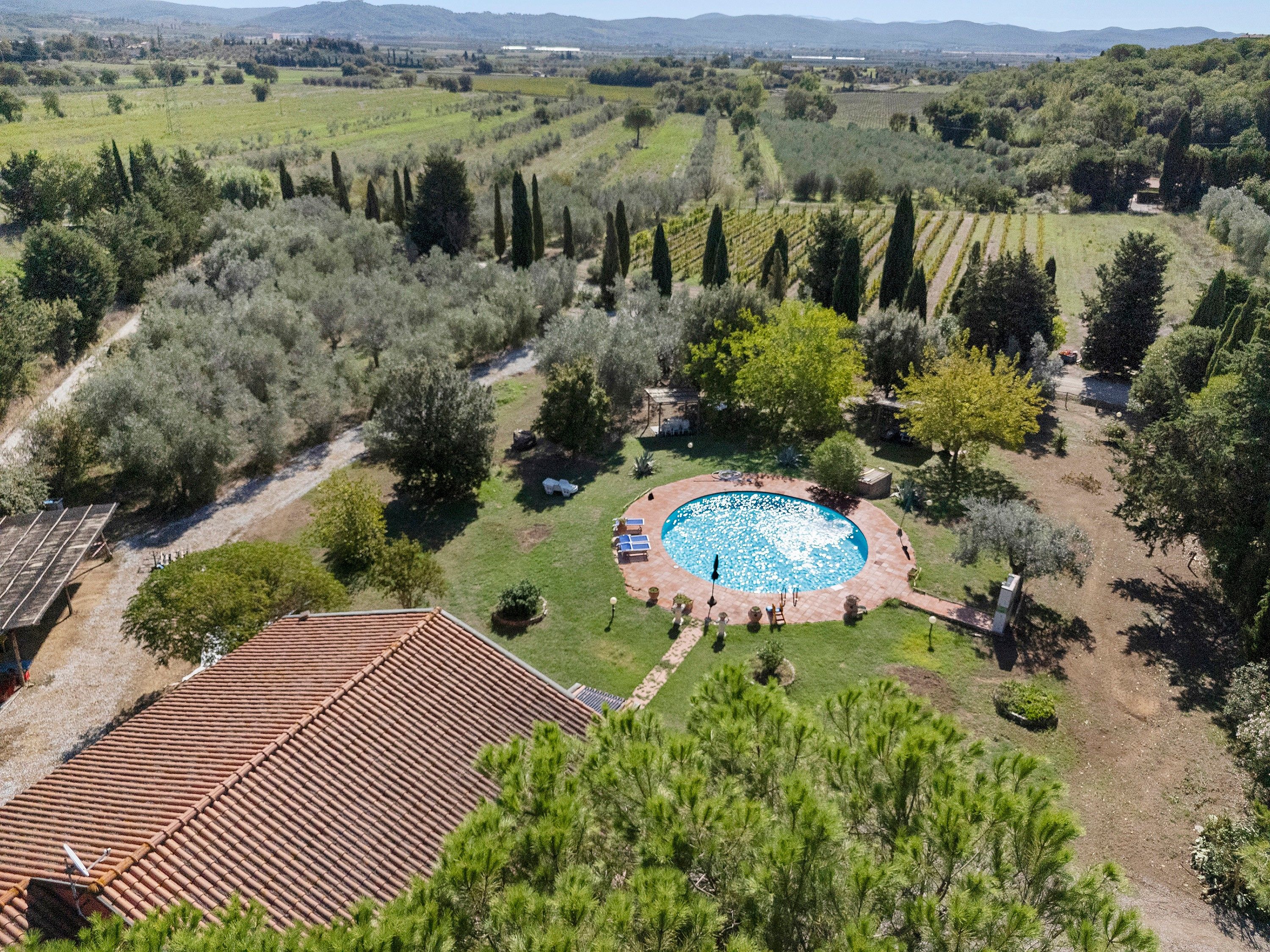 A residential property with a swimming pool surrounded by a garden, a tiled roof, and a view of the countryside.