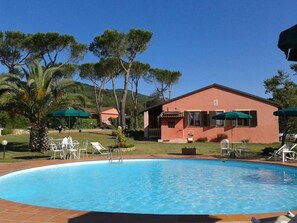 A swimming pool with lounge chairs and umbrellas, surrounded by trees and a building with a red facade.