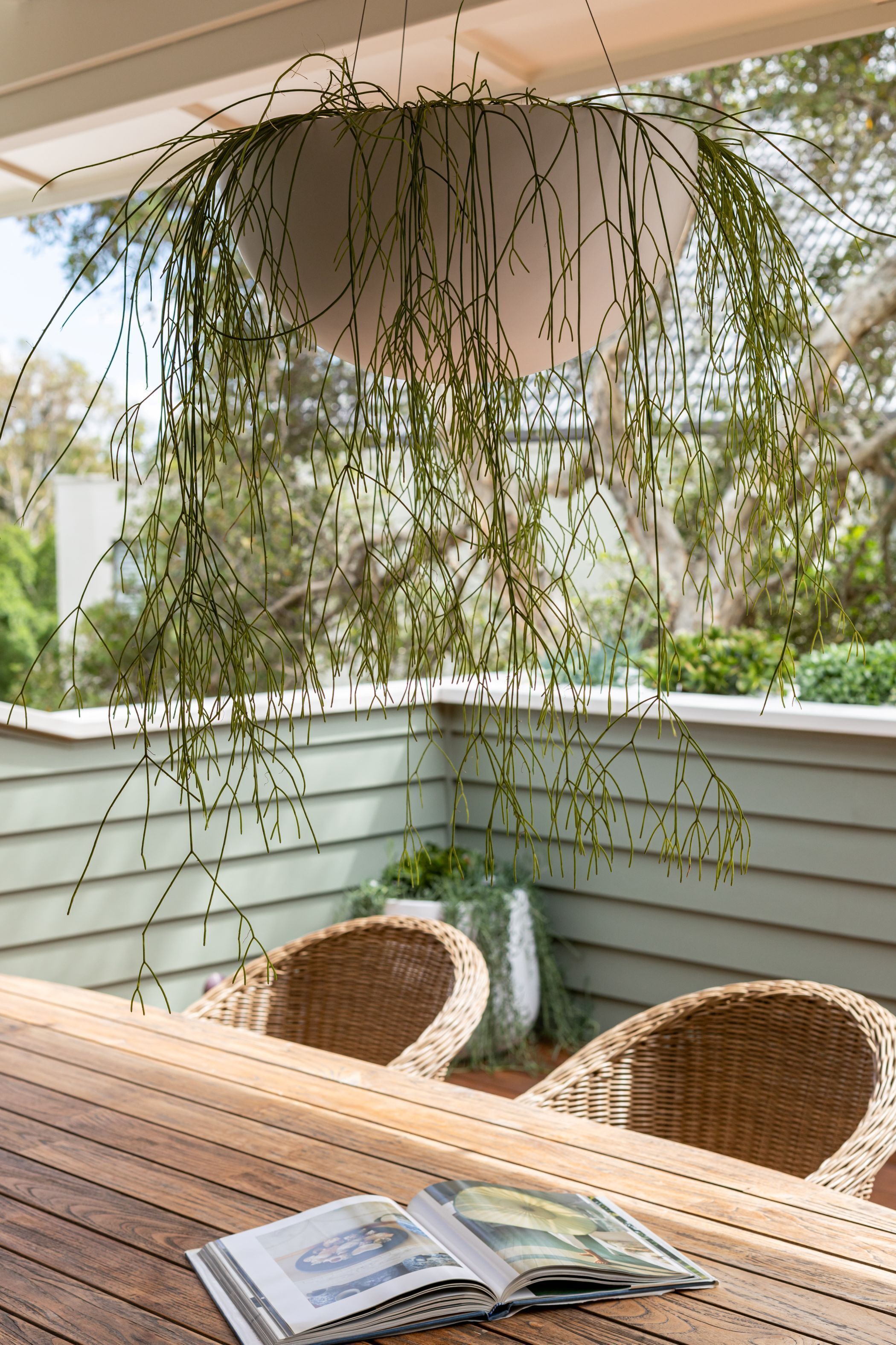 Une table en bois avec un magazine, des chaises en osier et des plantes suspendues dans un espace extérieur abrité.