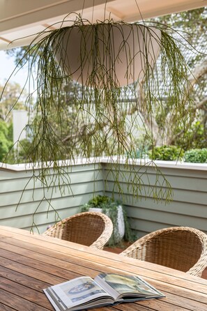 Una mesa de madera con una revista, sillas de mimbre y plantas colgantes en un espacio exterior cubierto.