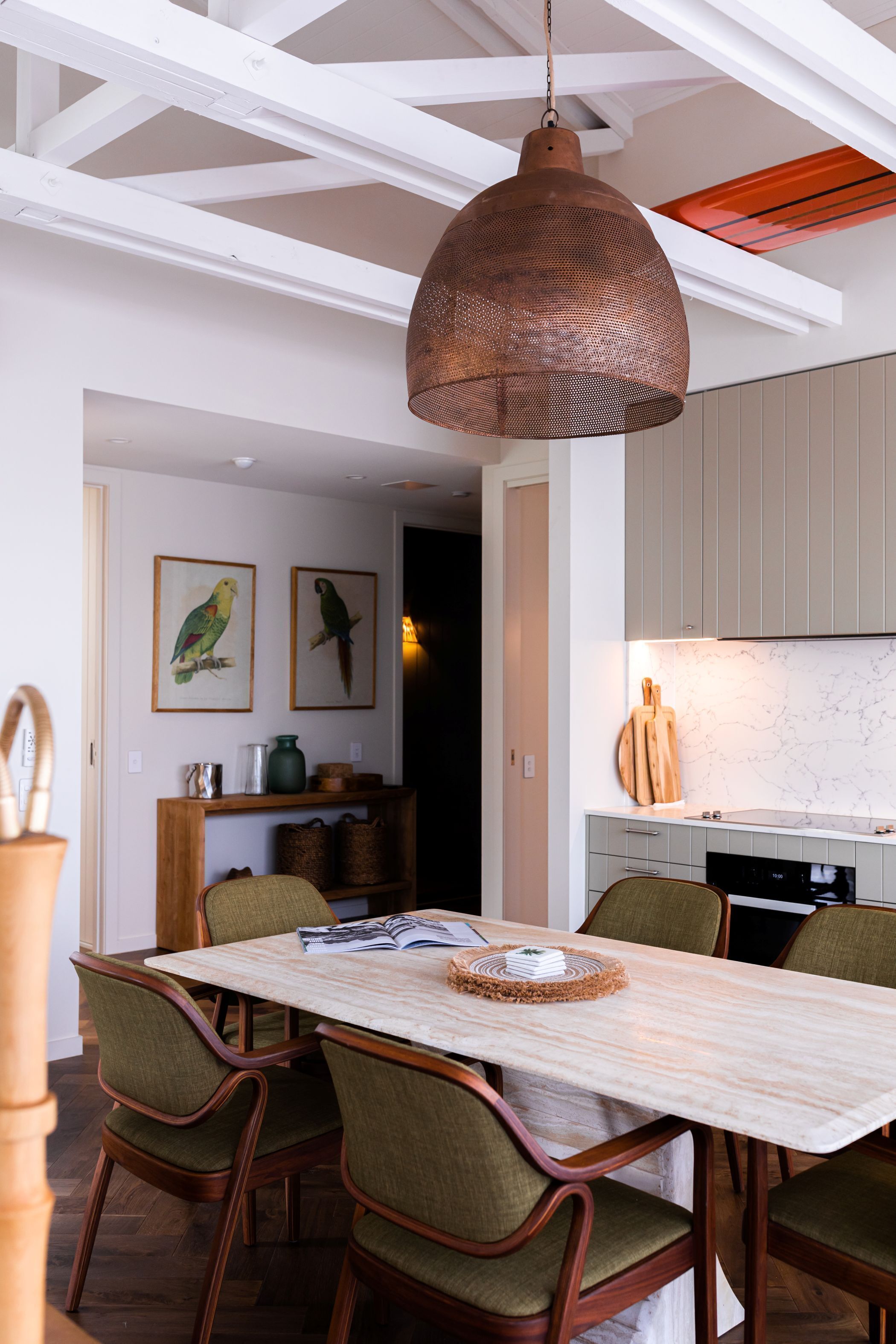A dining area with a wooden table and chairs, a hanging copper pendant light, and a kitchen in the background.