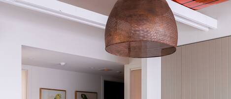A dining area with a wooden table and chairs, a hanging copper pendant light, and a kitchen in the background.