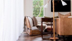A room with a wooden floor, a bed with a brown headboard, a TV mounted on the wall, and a window with blinds. - Sunbird Suite by Stay Noosa (Noosa Heads)