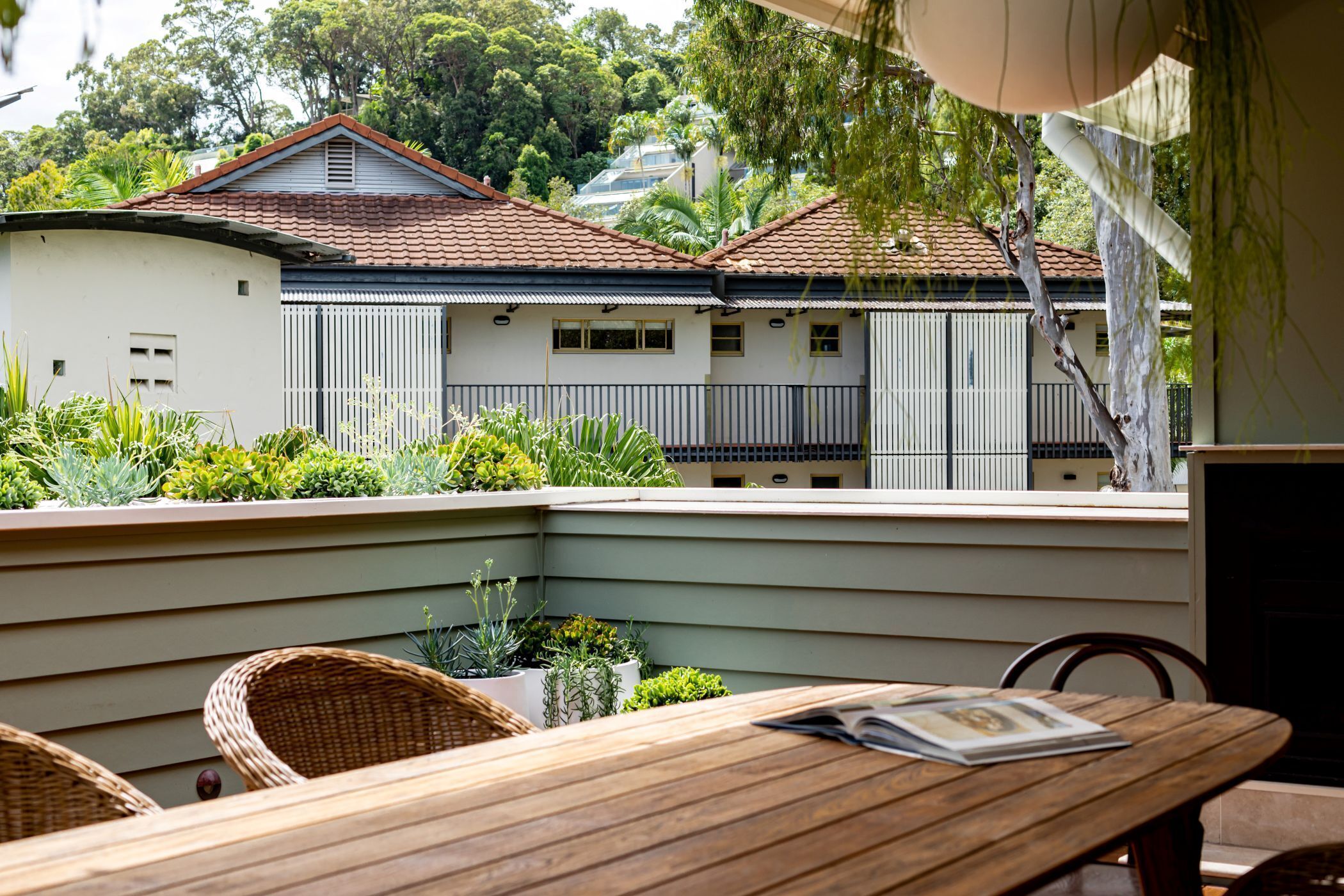 A wooden table with chairs and a magazine, a planter with plants, and a view of a building with a tiled roof.
