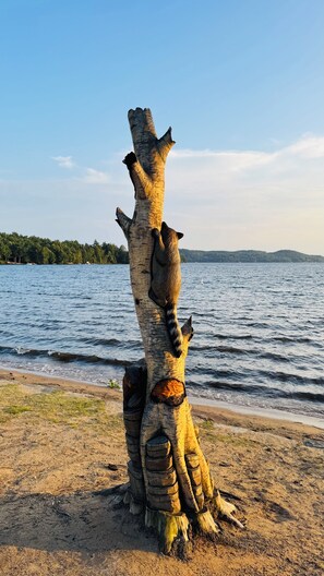 Eine Baumskulptur mit Tierfiguren an einem Strand, im Hintergrund ein See und ein Wald.