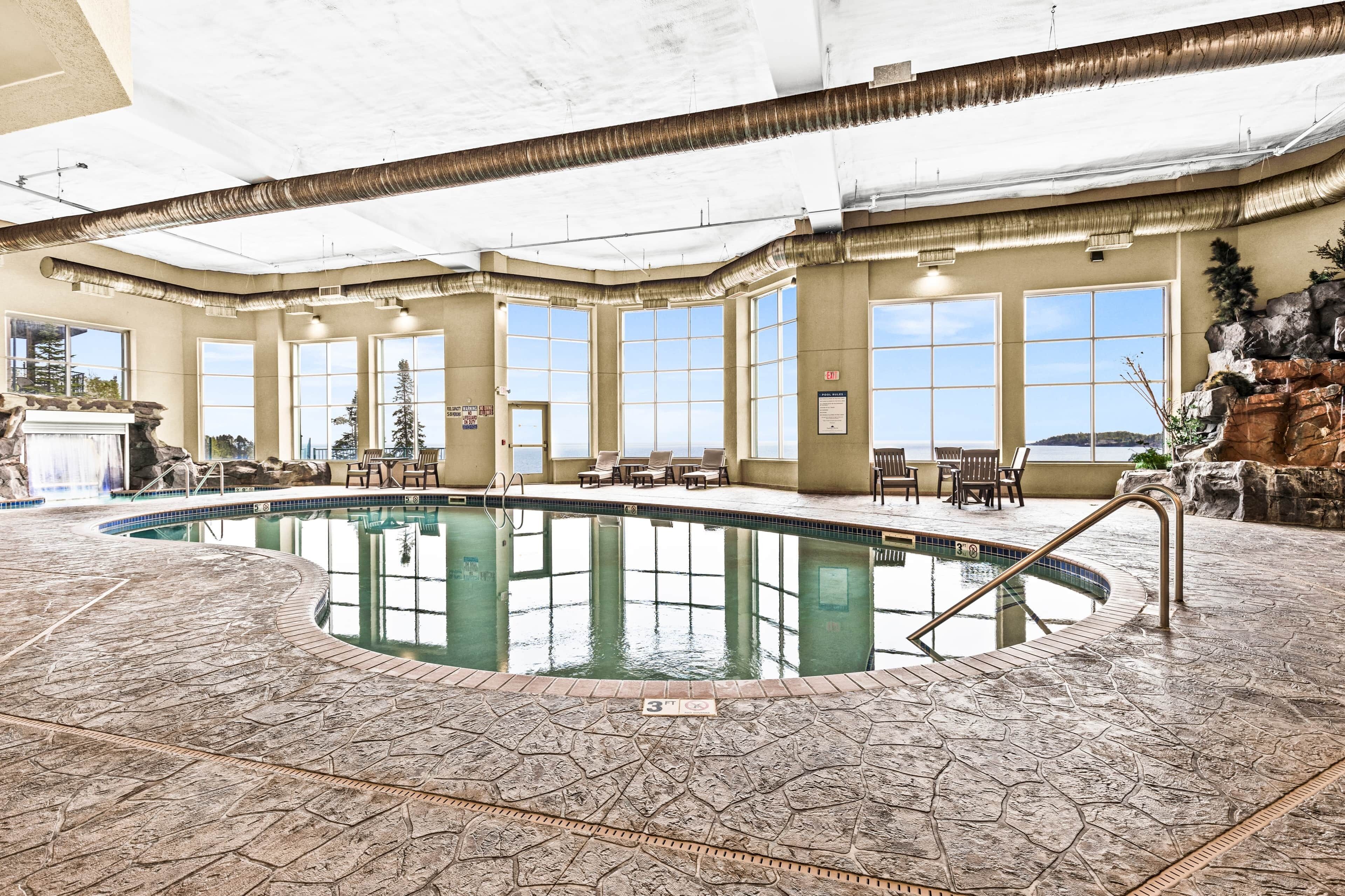 An indoor swimming pool with a stone floor, large windows, and a view of the outdoors.