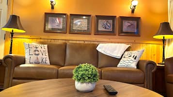 A room with a brown leather sofa, a round wooden table, a potted plant, and a remote control.