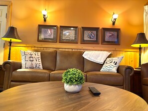 Een kamer met een bruine leren bank, een ronde houten tafel, een kamerplant en een afstandsbediening.