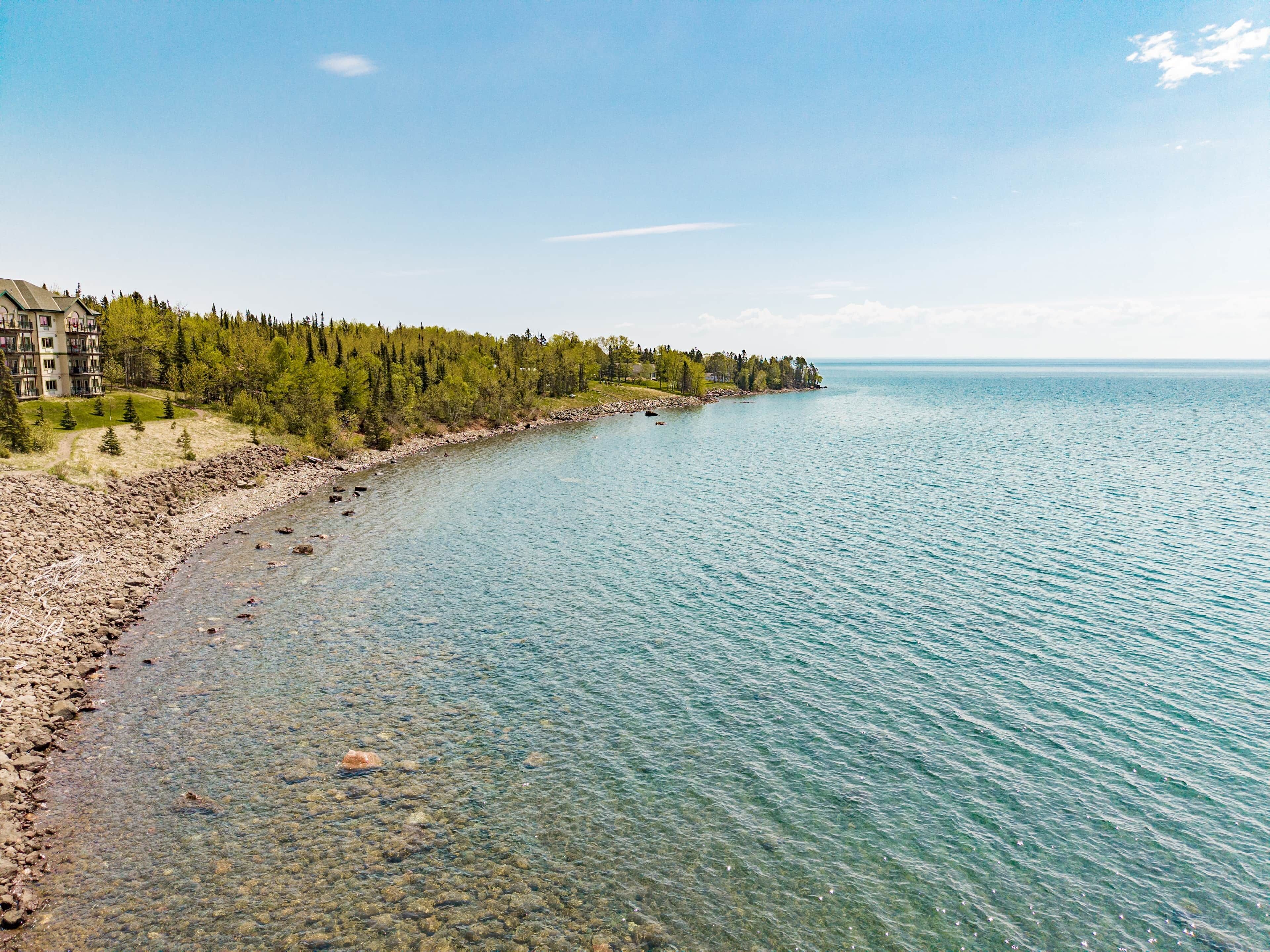 A coastal landscape with a clear shoreline, a rocky beach, and a forested area with a building on the left.