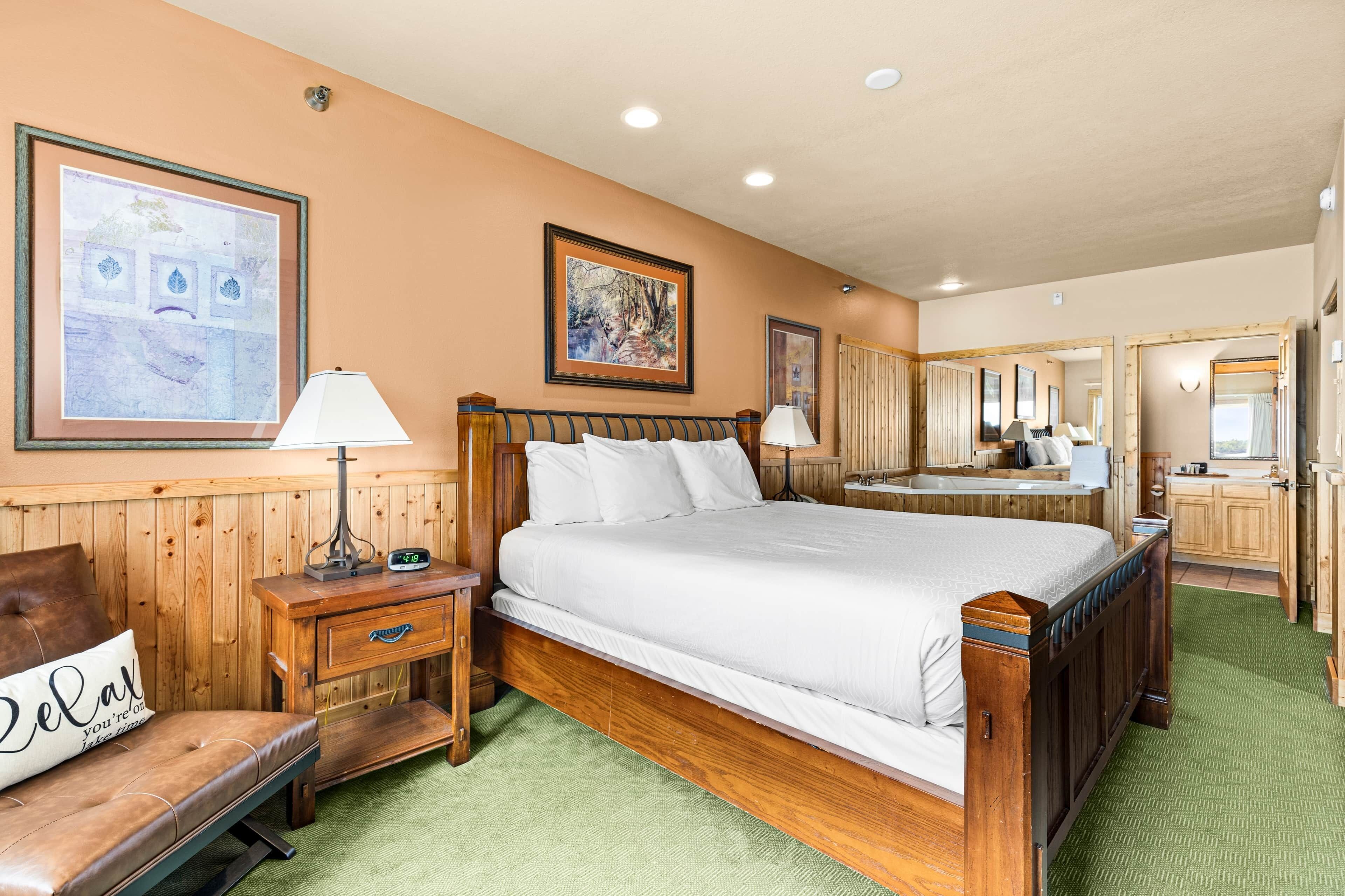 A hotel room with a large bed, a wooden nightstand, a bench with a pillow, and a bathroom visible through an open door.