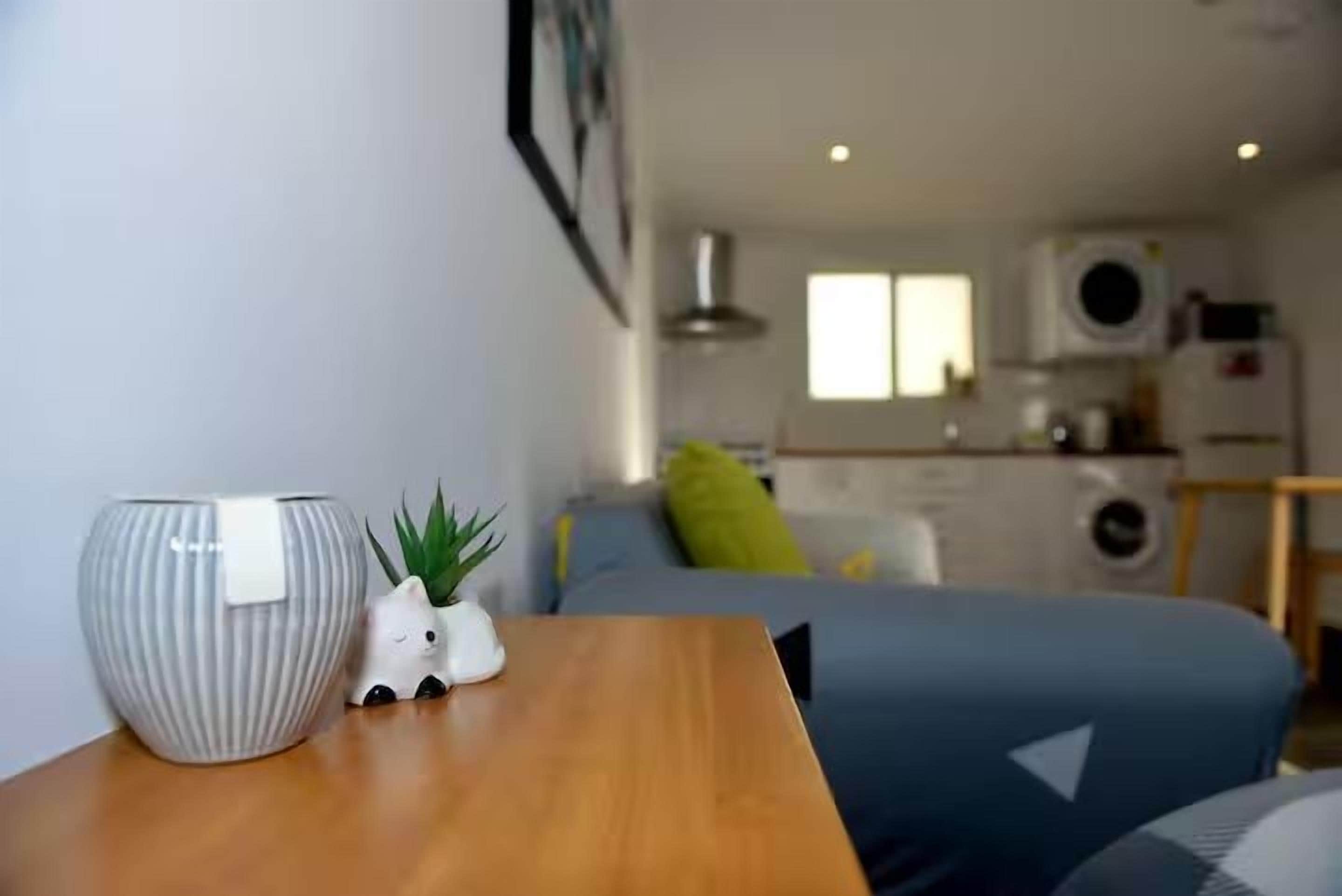 A compact living space with a bed, a small table with a vase and a plant, and a kitchenette in the background.