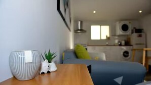 Un espace de vie compact comprenant un lit, une petite table avec un vase et une plante, et un coin cuisine à l’arrière-plan.