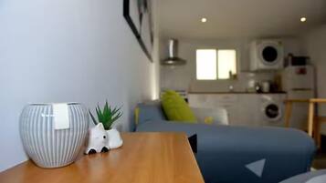 A compact living space with a bed, a small table with a vase and a plant, and a kitchenette in the background.