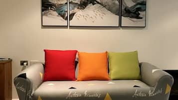 A grey sofa with three cushions in red, orange, and green colors, and a wall with three abstract paintings above it.