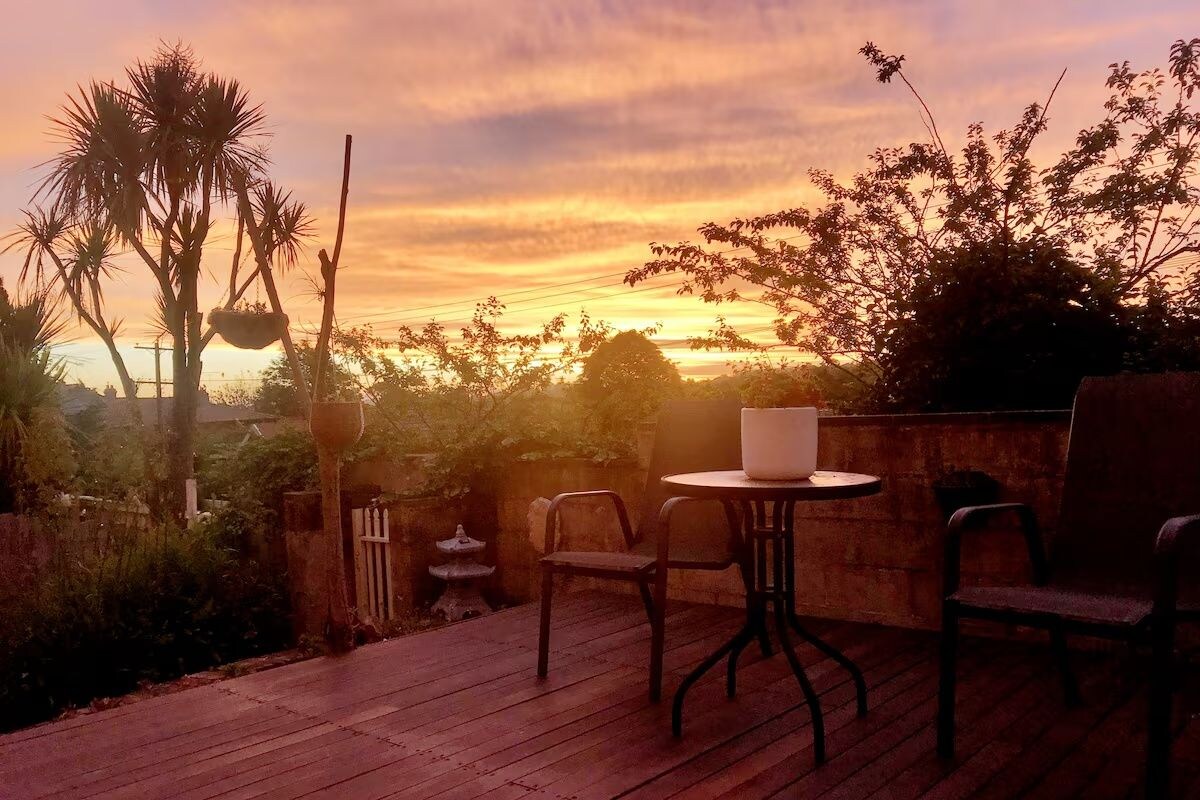 Un patio al aire libre con mesa y sillas, rodeado de vegetación y vistas al atardecer.