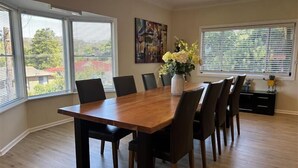 A dining room with a wooden table and chairs, large windows with blinds, a painting on the wall, and a vase with flowers on the table. - Cozy Mountain Cottage (Katoomba)