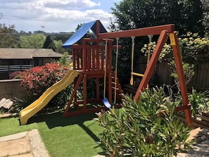 A wooden swing set with a slide, surrounded by green plants and a clear sky. - Cozy Mountain Cottage (Katoomba)
