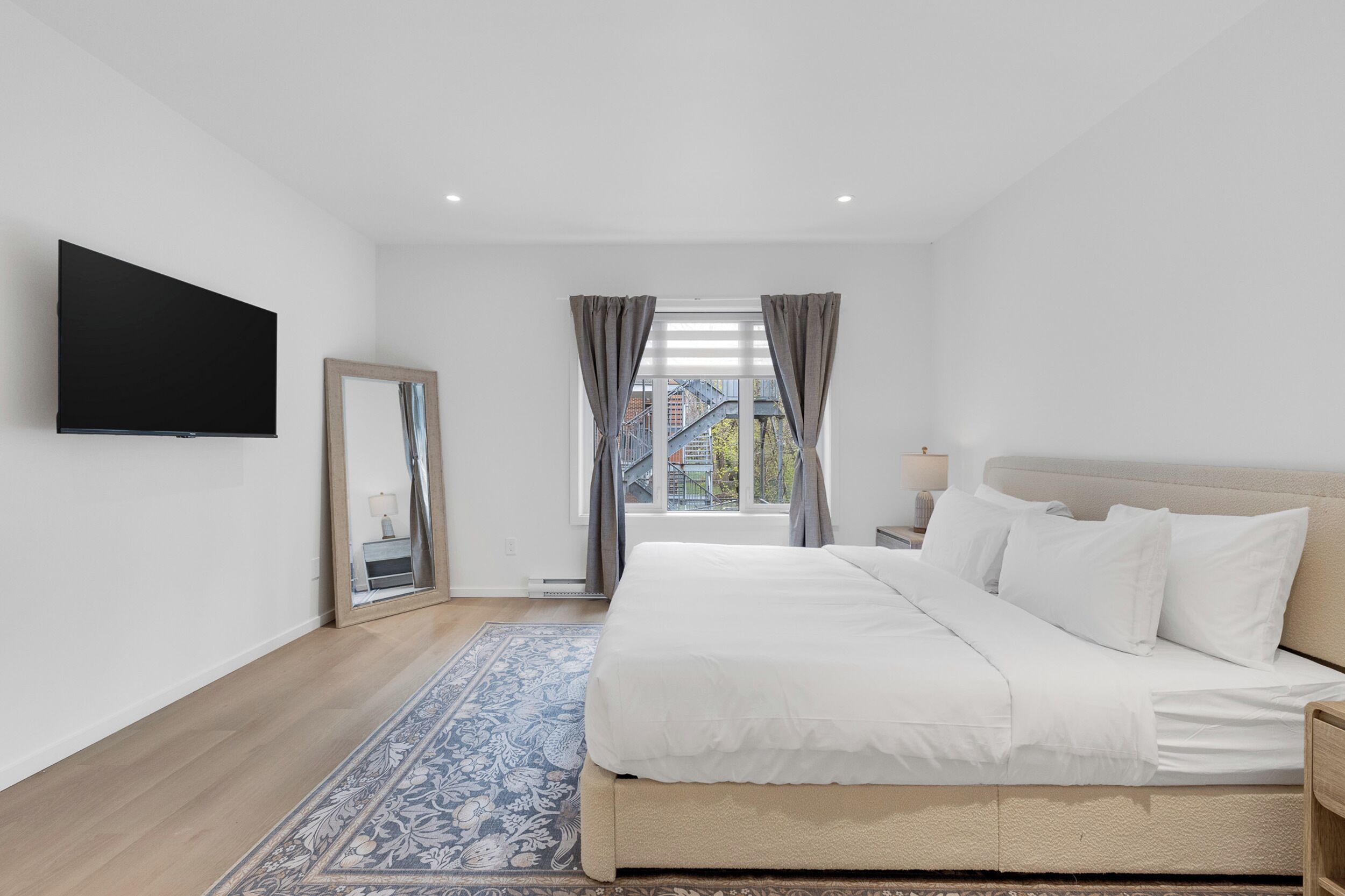 A bedroom with a bed, a TV mounted on the wall, a window with curtains, and a view of buildings outside.