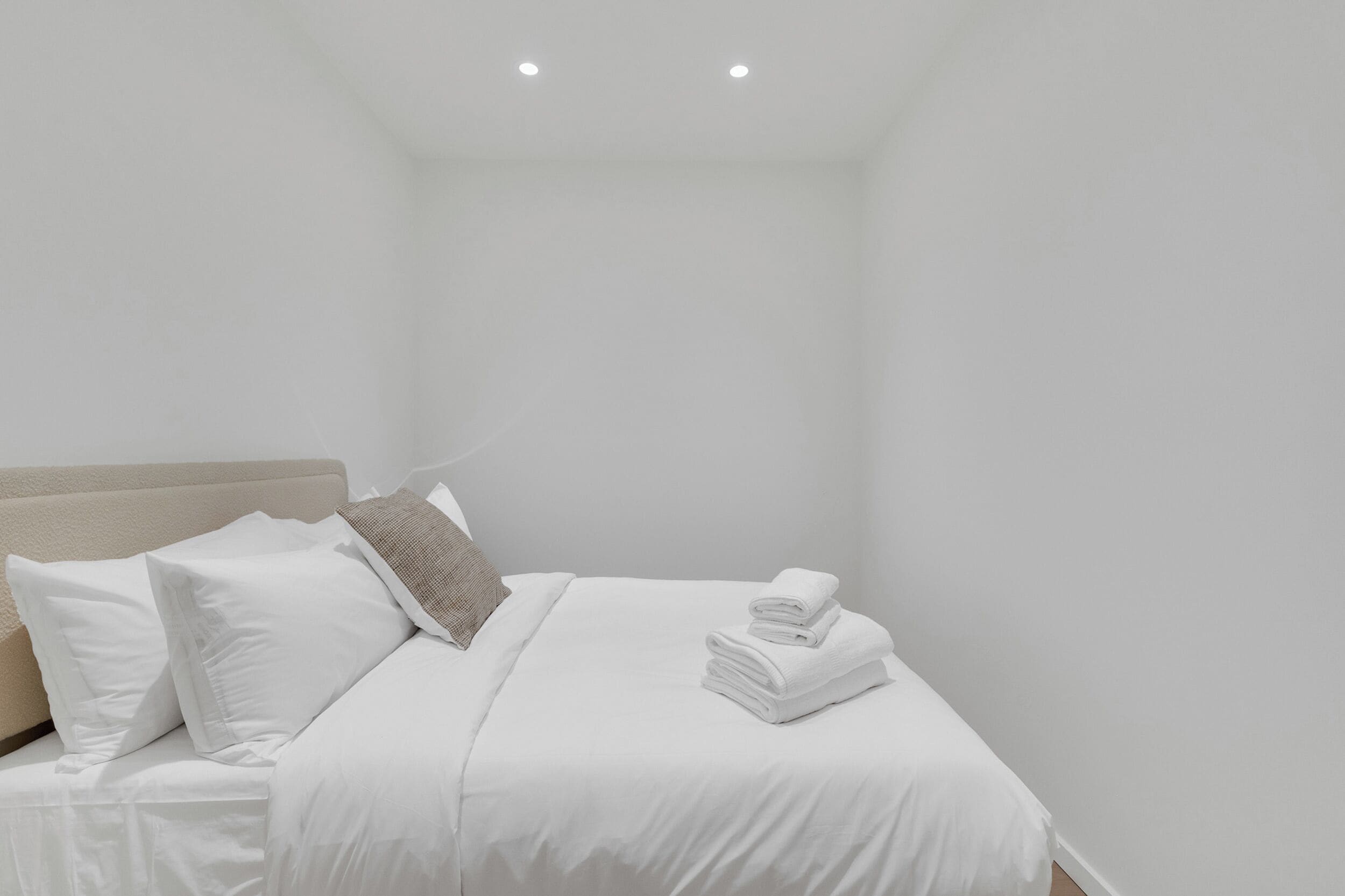 A neatly made bed with white linens and a folded towel, a beige headboard, and a single light fixture on the ceiling.